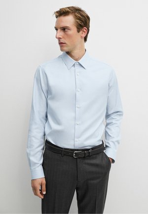 Ung mand iført en lyseblå langærmet skjorte med knapper, mørke bukser med striber, sort bælte, kigger mod venstre, foran en ensfarvet hvid baggrund.