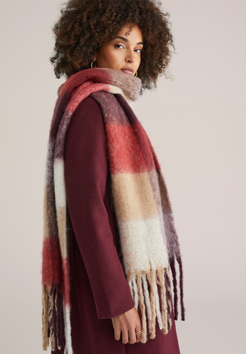 Sciarpa multicolore realizzata in tessuto morbido, con strisce rosse, beige e viola e dettagli con frange. Indossata sopra un cappotto bordeaux.
