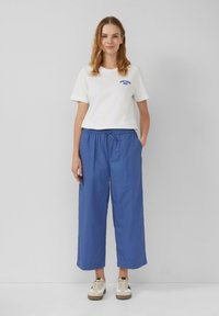 Maglietta bianca a maniche corte con testo blu e pantaloni ampi blu dal taglio rilassato. Le sneakers presentano un design beige e nero. Stile minimalista.