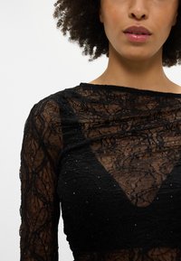 Čierny čipkový top s dizajnom do V, dlhými rukávmi a zložitými vzormi. Ozdobený malými trblietavými perličkami pre väčší detail.