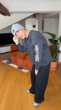 Sort langærmet skjorte med teksten "WORLD FAMOUS", kombineret med sorte bukser og sølvfarvede sko. Grå hue, indendørs med trægulv og planter.