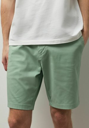 Mann trägt salbeigrüne knielange Shorts und ein weißes T-Shirt, Hände in den Taschen, steht vor einem neutralen Hintergrund.