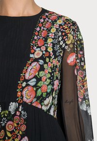 Schwarzes Kleid mit buntem Blumenstickerei am Oberteil, mit durchsichtigen langen Ärmeln und gefälteltem Detail. Weicher Stoff und lebendige Muster.