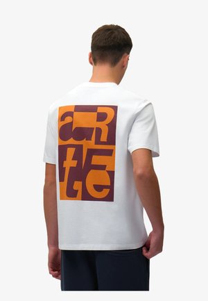 Uomo che indossa una maglietta bianca con grandi lettere astratte arancioni e marroni sul retro, in piedi contro uno sfondo chiaro e uniforme.