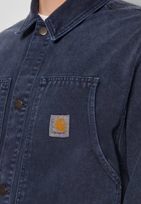 Námořnická džínová bunda s špičatým límcem, patentkami a přední kapsou se šitým logem Carhartt. Texturovaná tkanina.