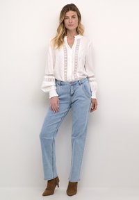 Camicia bianca con rifiniture in pizzo e chiusura a bottoni, abbinata a jeans a vita alta di colore azzurro chiaro. Completata con stivali con tacco in camoscio marrone.