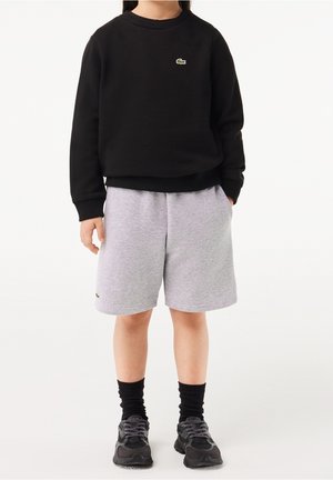 Felpa nera con scollo rotondo e logo verde, abbinata a pantaloncini grigi. Include tasche e scarpe sportive nere con calzini scuri.