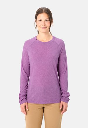 Vrouw met een paarse lange mouwen shirt en beige broek, rechtopstaand en glimlachend tegen een effen lichte achtergrond.