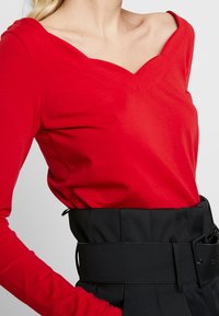Rotes Langarmoberteil mit weitem V-Ausschnitt, aus weichem Stoff gefertigt. Kombiniert mit einem schwarzen High-Waist-Gürtel und maßgeschneiderten Hosen.