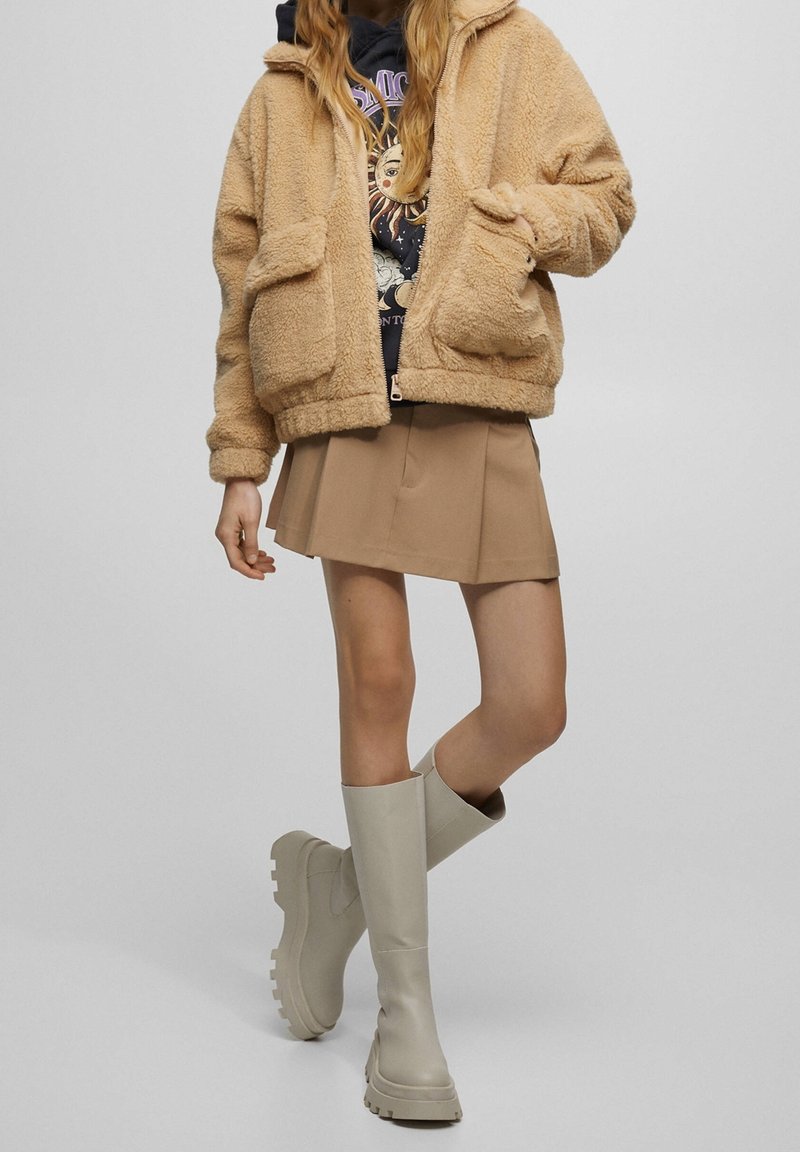 Veste en fausse fourrure beige avec capuche et deux poches avant, associée à une jupe plissée beige et des bottes en caoutchouc gris clair jusqu'aux genoux.