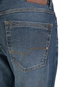 Gros plan sur la poche arrière d'un jean en denim bleu avec des coutures orange et une petite étiquette rouge, blanche et bleue sur la couture latérale.