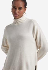 Cremefarbener Rollkragenpullover mit geripptem Kragen, lockerem Schnitt, weicher Textur, seitlichen Schlitzen und minimalen Nahtdetails.