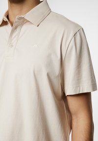 Kortärmad beige pikétröja i mjuk bomull med krage, knäppning framtill och en liten vit logotyp på bröstet. Slät textur.