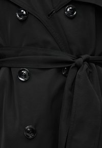 Trench-coat noir avec boutons noirs brillants et ceinture nouée à la taille, en vue rapprochée.
