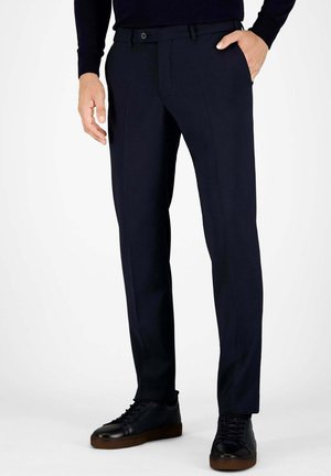 Mann trägt eng anliegende schwarze Hose und schwarze Ledersneaker, steht mit einer Hand in der Tasche vor einfarbigem Hintergrund.