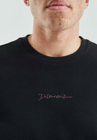 Schwarzes Baumwoll-T-Shirt mit kurzen Ärmeln und Rundhalsausschnitt. In hellrosa ist der Text "Sehe ich aus, als würde es mich interessieren?" nahe dem Saum gestickt.