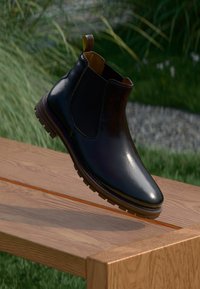 Botín Chelsea de cuero negro con un acabado brillante, paneles laterales elásticos y una suela de goma texturizada, exhibido sobre una superficie de madera.