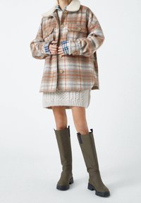 Giacca di lana a quadri in tonalità beige e ruggine con collo di pelliccia, abbinata a una gonna di maglia crema e stivali alti fino al ginocchio verde oliva.