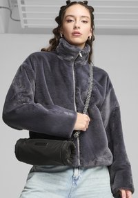 Giacca in pelliccia sintetica grigia con colletto alto e chiusura con zip, abbinata a una borsa cilindrica nera con tracolla intrecciata. Denim azzurro chiaro visibile.