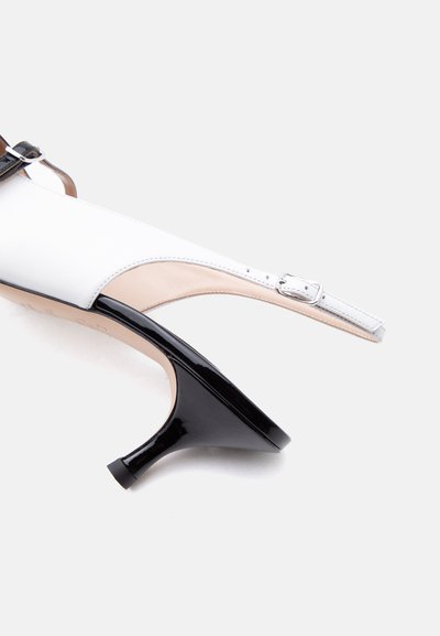 Λευκό παπούτσι με λουρί στην πλάτη, με μαύρο γυαλιστερό τακούνι στυλ kitten και ρυθμιζόμενο λουράκι με αγκράφα σε ανοιχτό φόντο.