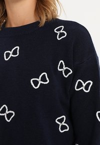 Marinblå tröja med vita broderade rosetter och dekorativa pärlor. Tyget verkar mjukt med ribbad halsringning och avslappnad passform.