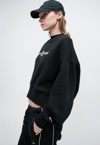 Černý zkrácený svetr s oversized rukávy, s bílým logem. Spárováno s černými tepláky s bílými akcenty. Čepice je také černá.