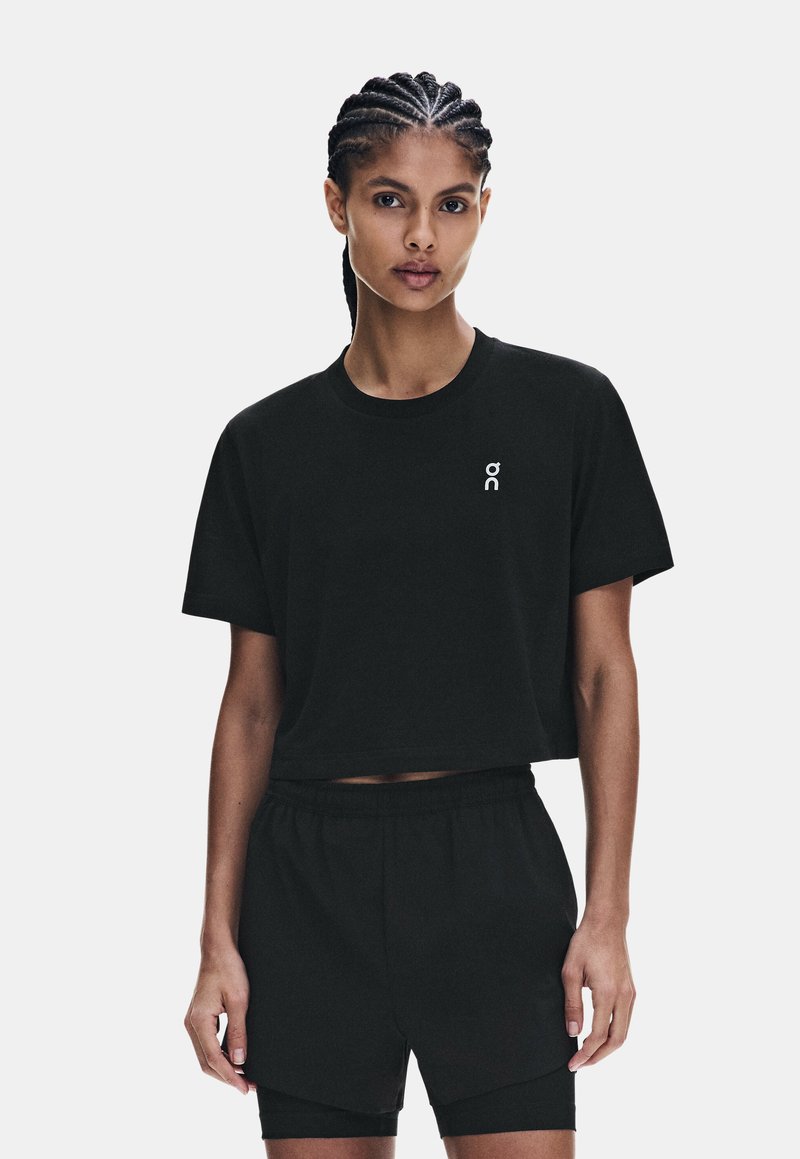 Femme aux cheveux tressés portant un t-shirt noir court et un short noir à plusieurs couches assorti, debout devant un fond clair uni.