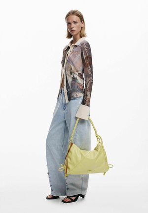 Gelbe Ledertasche mit einem geflochtenen Tragegurt, weicher Form und seitlichen Bändern. Das Model trägt eine gemusterte Bluse und weit geschnittene Jeans.