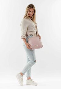 Borsa a tracolla in pelle rosa chiaro con una forma strutturata, hardware in argento e una lunga tracolla. Indossata da una persona in jeans chiaro e sneakers bianche.
