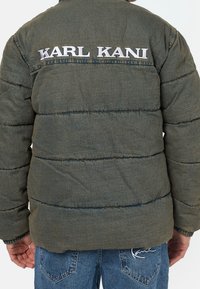 Kaki pufferjacka med horisontell sömnad och broderad "KARL KANI"-text på baksidan. Mjuk textur med hållbart material.