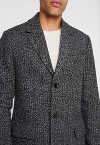 Blazer grigio testurizzato con un motivo sottile, colletto con risvolto, due bottoni e tasche laterali, indossato sopra un maglione color crema.