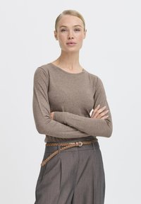 Žena s blond vlasmi, oblečená v hnedom topu s dlhým rukávom a šedých nohaviciach s vysokým pásom, s hnedým pletencovým opaskom, so založenými rukami a neutrálnym výrazom.