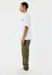 Witte T-shirt met korte mouwen en zwart logo, olijfgroene cargobroek met meerdere zakken, en witte sneakers. Eenvoudig ontwerp, casual pasvorm.