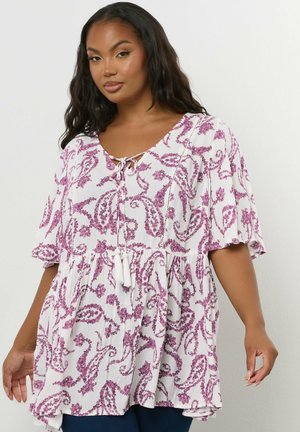 Femme portant un chemisier ample à motif cachemire blanc et violet, avec un lien au niveau de l'encolure et de larges manches courtes.