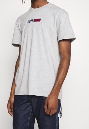 Mand iført lysegrå Tommy Jeans t-shirt med logopatch på bryst og ærme, kombineret med mørkeblå jeans med hvide syninger.