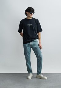 Μαύρο κοντομάνικο T-shirt με τυπωμένο λογότυπο, ανοιχτού μπλε ξεθωριασμένο τζιν και λευκά αθλητικά παπούτσια. Το μοντέλο φοράει σκούρα γυαλιά ηλίου, στέκεται σε γκρι δάπεδο.