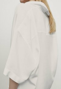 Biała, oversize'owa koszula z kapturem, posiadająca krótkie rękawy, gładką fakturę i luźny krój. Minimalistyczny design, bez wyraźnych wzorów.