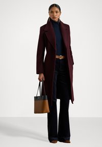 Soprabito in lana bordeaux con rever a punta, abbinato a un dolcevita blu navy e pantaloni scuri. Tiene una borsa bicolore, con sezioni nere e marroni.