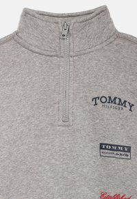 Γκρίζα φλις ζακέτα με ψηλό γιακά, μισό φερμουάρ και ναυτικό εκτυπωμένο λογότυπο στο στήθος. Διαθέτει ραμμένη κόκκινη λεπτομέρεια "Iδρύθηκε".