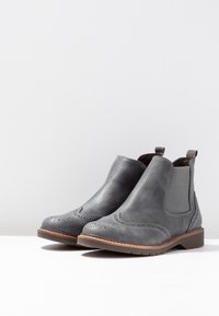 Šedé kožené kotníkové boty s brogue detaily, elastickými bočními panely a kontrastní hnědou gumovou podrážkou. Hladký povrch a kulatá špička.