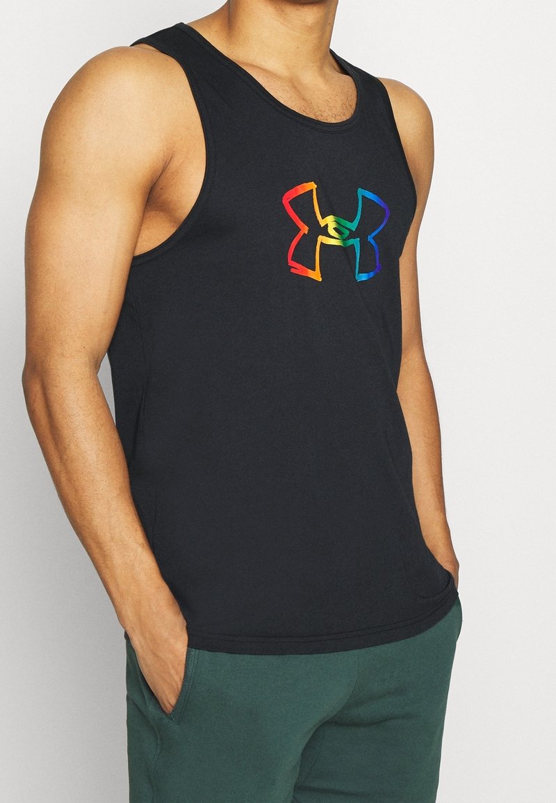 Homme portant un débardeur noir avec un logo Under Armour aux couleurs de l'arc-en-ciel et un pantalon vert, les mains dans les poches.