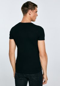 Jonge man van achteren gezien, gekleed in een strakke zwarte korte mouw T-shirt en donkere spijkerbroek, tegen een lichte effen achtergrond.