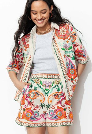 Femme souriante aux longs cheveux foncés, portant une chemise à motif floral coloré et un short assorti par-dessus un haut gris clair uni.