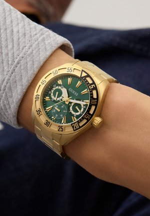 Goldene Metallarmbanduhr mit grünem Zifferblatt, drei Unterzifferblättern und schwarz-goldener Lünette, getragen am linken Handgelenk über einem hellgrauen Langarmshirt.