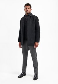 Zwarte wollen blendjas met een hoge kraag, gelaagd over een zwarte pufferjack. Grijze broeken en zwarte enkellaarsjes maken de look compleet.