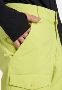 Gele cargobroek met een gladde textuur, voorzien van grote zakken, een drukknoop en een rits, gecombineerd met een zwart shirt met lange mouwen.