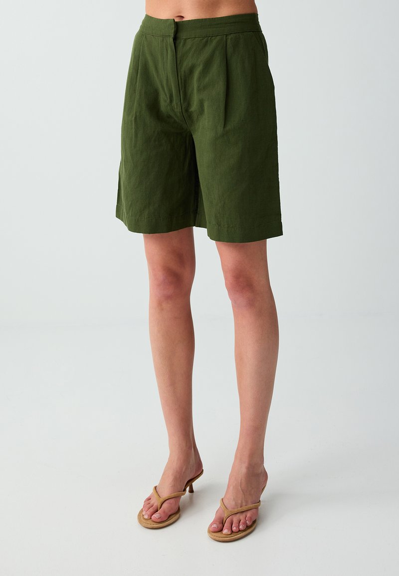 Femme portant un short kaki arrivant aux genoux et des sandales beiges à talons, debout sur un fond clair uni.