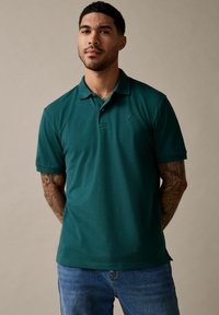 Polo shirt de manga corta verde oscuro con cuello, que presenta un logo sutil en el pecho y puños acanalados, combinado con jeans azules.
