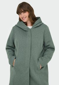 Manteau à capuche vert en tissu texturé, doté d'une fermeture éclair à l'avant et de poches latérales, conçu avec une coupe décontractée et un style minimaliste.