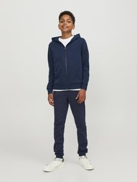 Sudadera con cremallera color marino con bolsillos frontales, combinada con pantalones jogger color marino y zapatillas blancas con detalles verdes. Tejido suave, ajuste relajado.